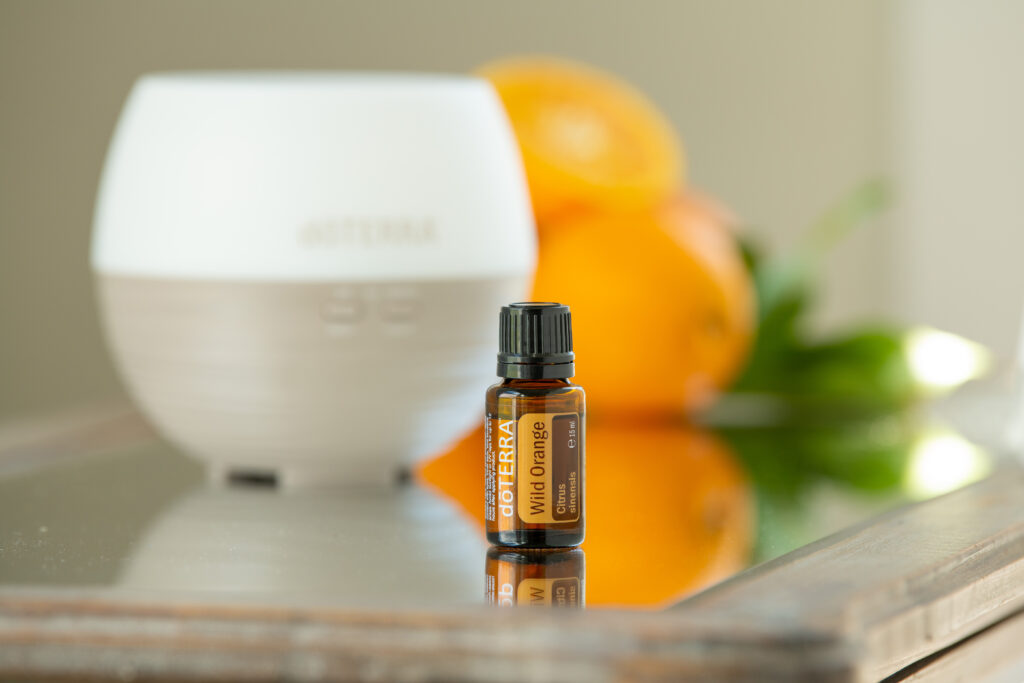 Olejek Wild Orange dōTERRA obok dyfuzora i świeżych owoców pomarańczy na stole.